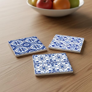 May include: Three square ceramic coasters with a blue and white floral pattern are arranged on a wooden surface. The coasters have a decorative design, and a bowl of fruit is in the background.