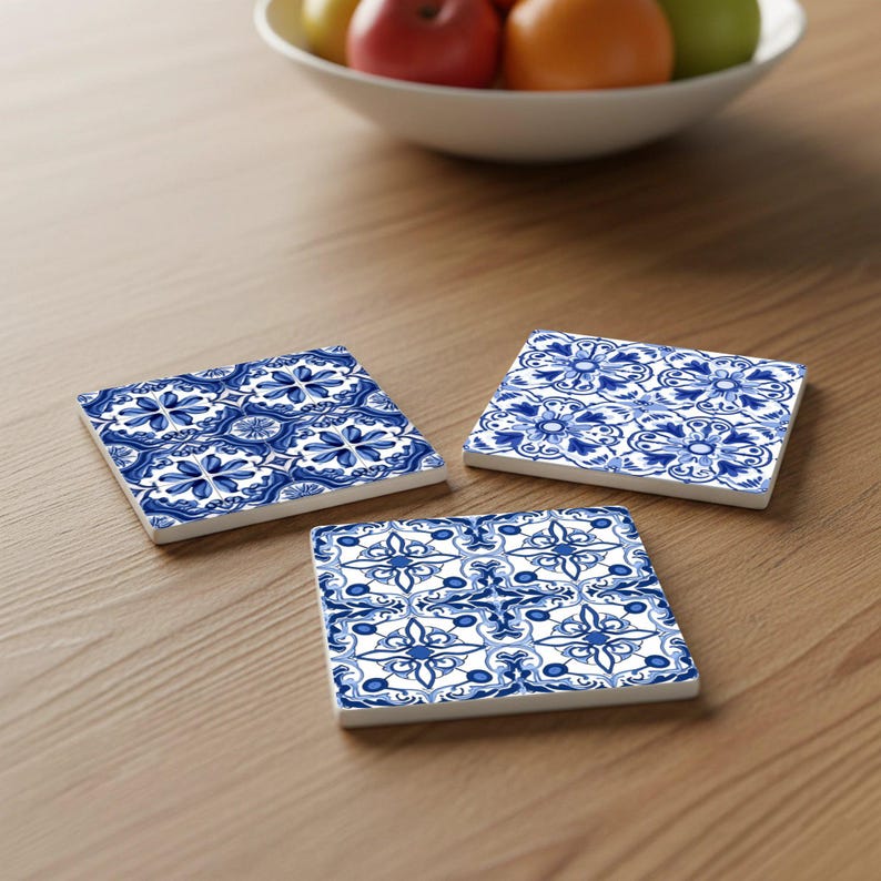 May include: Three square ceramic coasters with a blue and white floral pattern are arranged on a wooden surface. The coasters have a decorative design, and a bowl of fruit is in the background.
