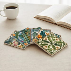 Op de afbeelding: Een verzameling vierkante keramische onderzetters met botanische ontwerpen, met verschillende tropische planten en bloemen in groen, blauw en oranje tinten. Een witte koffiekop en een open boek zijn op de achtergrond te zien.