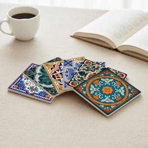 May include: A set of decorative square coasters featuring detailed floral and geometric designs in shades of blue, orange, green, and yellow. The coasters are displayed on a light surface, with a coffee cup and an open book visible in the background.