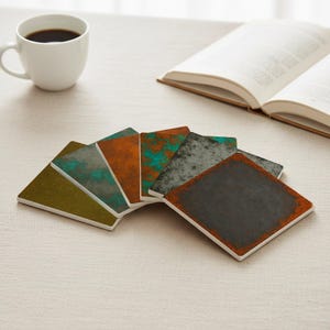 Op de afbeelding: Een verzameling vierkante onderzetters met een verweerde, getextureerde uitstraling. De onderzetters hebben verschillende kleuren, waaronder olijfgroen, blauwgroen, oranje en grijs, met witte randen. Een witte koffiekop en een open boek staan op de achtergrond.