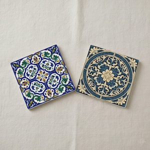 K&ouml;nnte beinhalten: Zwei quadratische Keramikuntersetzer mit aufwendigen Blumenmustern. Einer hat ein blaues, gr&uuml;nes und gelbes Design auf wei&szlig;em Grund. Der andere hat ein kreisf&ouml;rmiges Muster in Blau und Creme. Dekorative Artikel, ca. 10 cm x 10 cm.