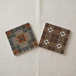 May include: Two square coasters with a steampunk design. One coaster features a pattern of squares in shades of gray, brown, and copper, with metal rivet details. The other coaster has a similar design with white and brown accents.
