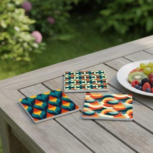 May include: Three square ceramic coasters with retro geometric patterns in teal, orange, yellow, and cream colours. The coasters are arranged on a wooden table, with a plate of fruit in the background. The coasters are approximately 10 cm square.