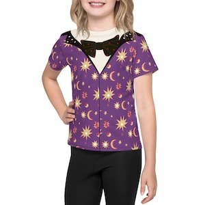 May include: A purple t-shirt featuring a whimsical design of a tuxedo with a black bow tie and a pattern of gold stars, moons, and red accents. The shirt has a cream-colored collar and is worn by a person.