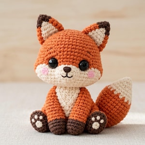 Könnte beinhalten: Ein gehäkeltes Fuchskuscheltier aus orangefarbenem Garn mit cremefarbener Brust, Schnauze und Schwanzspitze. Es hat große schwarze Kunststoffaugen, eine kleine braune Nase und rosa gestickte Wangen. Die Pfoten sind dunkelbraun mit cremefarbenen Ballen.