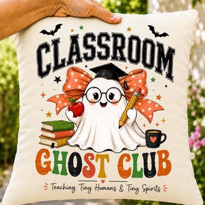 Può includere: Un cuscino decorativo color crema con l'illustrazione di un fantasma con occhiali e tocco da laureato, che tiene in mano una mela e una matita. Il testo recita CLASSROOM GHOST CLUB e Teaching Tiny Humans & Tiny Spirits, con libri e una tazza.