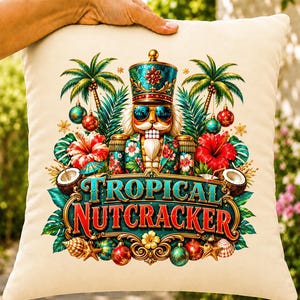 Può includere: Un cuscino decorativo color crema con la grafica di uno schiaccianoci che indossa occhiali da sole, circondato da palme, fiori di ibisco, noci di cocco e ornamenti. Il testo TROPICAL NUTCRACKER è stampato in grandi lettere stilizzate al centro.