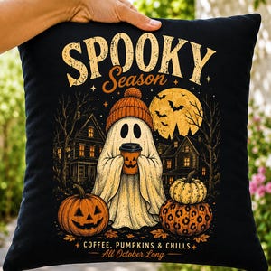 Puede incluir: Un cojín cuadrado negro con un gráfico de un fantasma con un gorro naranja que sostiene una taza de café con cara de calabaza. El diseño incluye el texto SPOOKY Season, COFFEE, PUMPKINS & CHILLS, All October Long, junto con calabazas, murciélagos y casas.