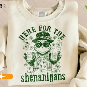 Op de afbeelding: Crèmekleurige sweatshirt met een groene afbeelding van een stripfiguur met een zonnebril, hoed en overalls, met twee bierpullen. De tekst "HERE FOR THE shenanigans" is groen gedrukt.