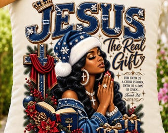 May include: A cream-colored throw pillow featuring a graphic of a person wearing a blue Santa hat, praying with hands together. The text reads JESUS The Real Gift and includes the Bible verse Isaiah 9:6, a cross, a Bible, and gifts.