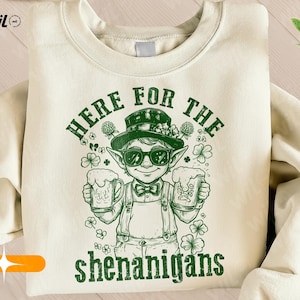 Puede incluir: Sudadera color crema con un gráfico verde de un duende con gafas de sol y sombrero, sosteniendo dos jarras de cerveza. El texto "HERE FOR THE shenanigans" está impreso encima y debajo de la ilustración.