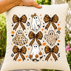 Puede incluir: Un cojín cuadrado de color blanco roto con un patrón repetitivo de fantasmas y lazos con estampado de leopardo. El diseño incluye fantasmas con patrones de leopardo, fantasmas con lazos y fantasmas con calabazas naranjas, acentuados por pequeñas estrellas y corazones naranjas.