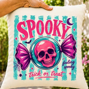 Puede incluir: Un cojín cuadrado de color blanco roto con un estampado gráfico que incluye los textos "SPOOKY", "feeling spooky" y "trick or treat". El diseño muestra una calavera rosa dentro de un envoltorio de caramelo azul, una luna creciente y una telaraña.