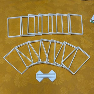 May include: A collection of white, square-shaped plastic frames, possibly for crafting or display, arranged on a yellow patterned surface. A small, white, bow-tie shaped piece is also present.
