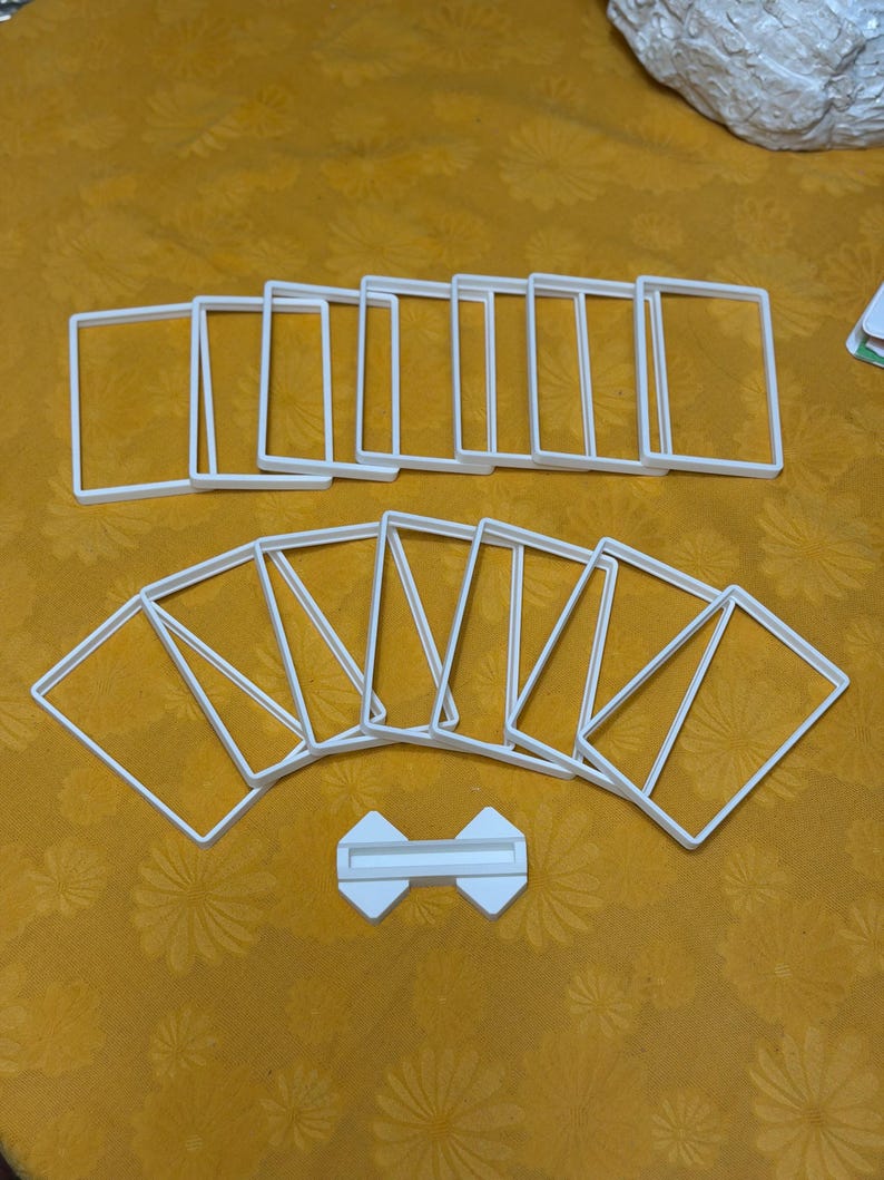 May include: A collection of white, square-shaped plastic frames, possibly for crafting or display, arranged on a yellow patterned surface. A small, white, bow-tie shaped piece is also present.