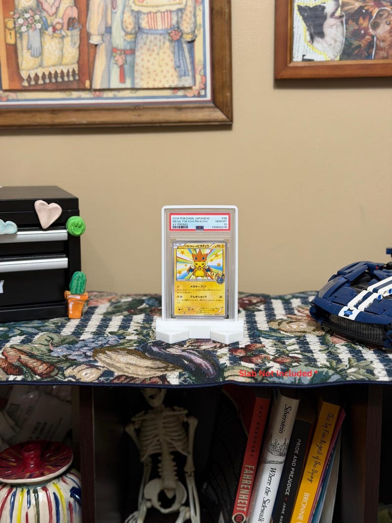 May include: A graded Pok&eacute;mon trading card featuring Pikachu, displayed in a clear plastic case. The card is held in a white stand on a floral patterned surface. Artwork and text are visible on the card. The background includes framed art and a small cabinet.