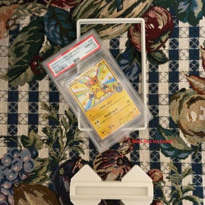 May include: A graded Pok&eacute;mon card in a protective case. The card features a Pikachu illustration with Japanese text. The case is labeled with details including the card's grade and a unique identification number. A white frame and a white display stand are also present.