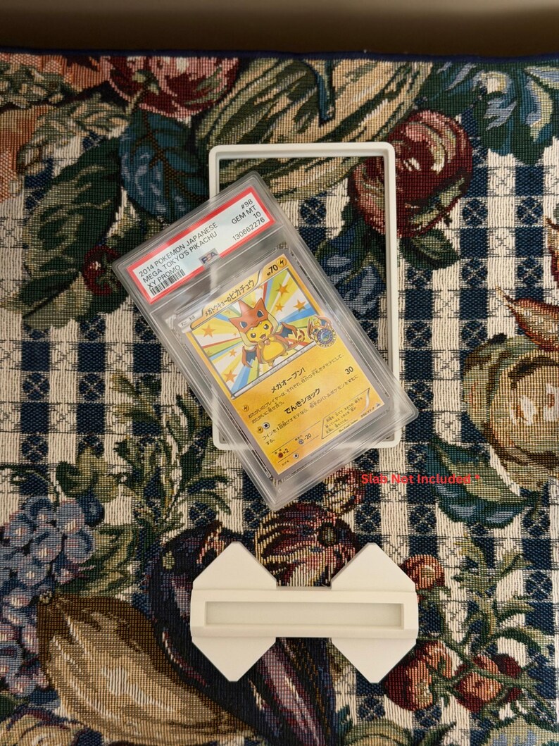 May include: A graded Pok&eacute;mon card in a protective case. The card features a Pikachu illustration with Japanese text. The case is labeled with details including the card's grade and a unique identification number. A white frame and a white display stand are also present.