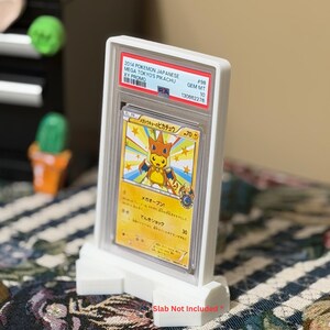 May include: A white display stand holding a Pokemon trading card. The card is encased in a protective holder with text that reads "2014 Pokemon Japanese Mega Tokyo's Pikachu XY Promo." The card features a yellow Pikachu character.