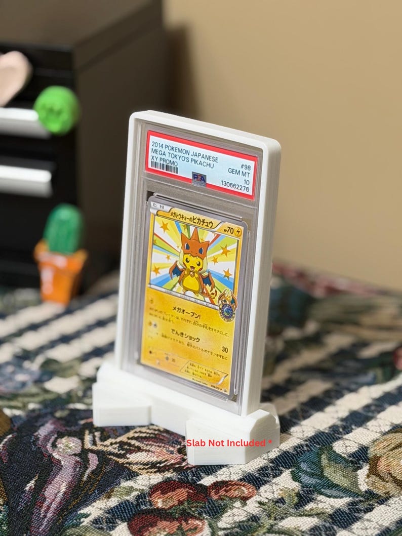 May include: A white display stand holding a Pokemon trading card. The card is encased in a protective holder with text that reads "2014 Pokemon Japanese Mega Tokyo's Pikachu XY Promo." The card features a yellow Pikachu character.