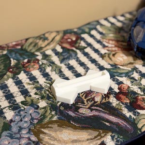 May include: A small, white, plastic object with an arrow-like shape. The object is resting on a patterned fabric with a blue and white checkered design, and floral and fruit motifs in shades of red, green, and purple.