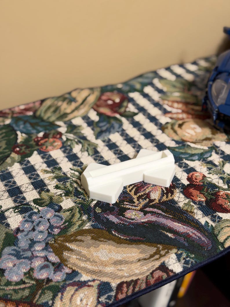 May include: A small, white, plastic object with an arrow-like shape. The object is resting on a patterned fabric with a blue and white checkered design, and floral and fruit motifs in shades of red, green, and purple.