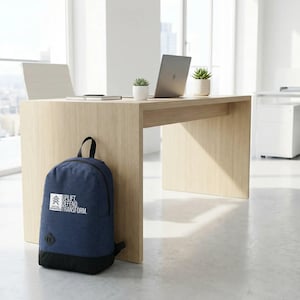 May include: A navy blue backpack with a black base rests against a light wood desk in a modern office setting. The backpack features the text "UPLIFT DEFEND TRANSFORM" in white. A laptop and small potted plants sit on the desk.
