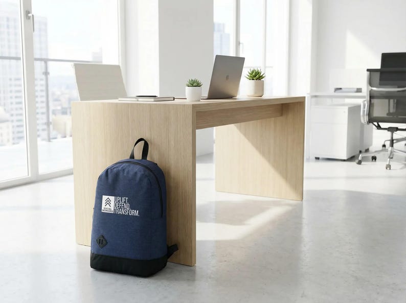 May include: A navy blue backpack with a black base rests against a light wood desk in a modern office setting. The backpack features the text "UPLIFT DEFEND TRANSFORM" in white. A laptop and small potted plants sit on the desk.
