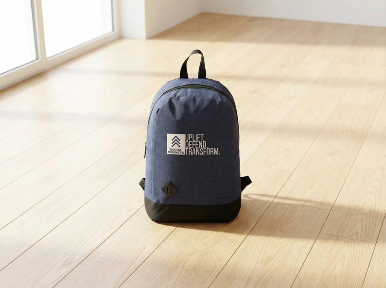 May include: A navy blue backpack with a black base and handle. The backpack features the text "UPLIFT. DEFEND. TRANSFORM." and the words "SOCIAL WORKERS" in a white square. The backpack is on a light wood floor.