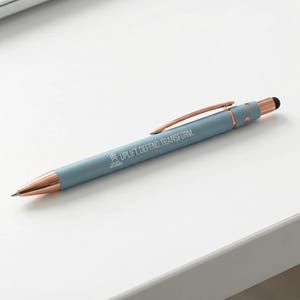 May include: A light blue pen with rose gold accents and a stylus tip. The pen has the words "UPLIFT. DEFEND. TRANSFORM." printed on the barrel. The pen is lying on a white surface.
