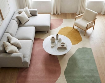 May include: An overhead shot of a living room with a light gray sectional sofa, a white armchair, and a round marble coffee table. A large area rug features abstract shapes in pink, yellow, and green. Decorative vases and books are on the table.