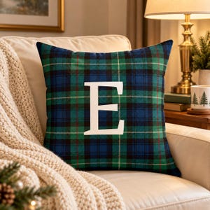 May include: A square throw pillow featuring a blue and green plaid pattern with thin red accent lines. A large, bold, white capital letter E is centered on the front of the pillow. The fabric has a soft, woven texture and a classic tartan design.