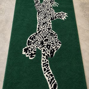 Op de afbeelding: Een rechthoekig groen tapijt met een zwart-wit luipaardontwerp. De luipaard wordt afgebeeld in een springende pose, met zijn vlekken en vorm die afsteken tegen de effen groene achtergrond. Het tapijt is waarschijnlijk gemaakt van een zacht, getextureerd materiaal.