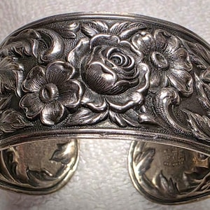 May include: A wide silver cuff bracelet featuring a high-relief repoussé design of blooming roses and intricate foliage. The metal surface has a polished finish with darkened recessed areas to highlight the detailed floral patterns and textures.
