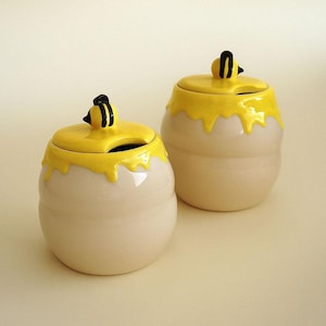 Può includere: Due vasetti di miele in ceramica con coperchi gialli e design a goccia giallo. Ogni coperchio ha una piccola figura di ape nera e gialla. I vasetti sono bianco sporco e hanno una forma arrotondata.