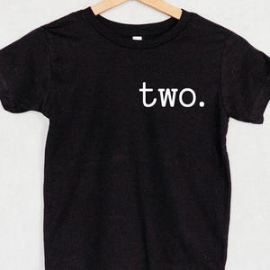 May include: A black short-sleeved t-shirt hanging on a wooden hanger. The shirt features the word two. printed in white lowercase letters on the upper right chest area. The garment has a crew neckline and a standard straight hemline.
