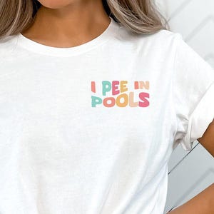 May include: White t-shirt with rolled-up sleeves and a knot at the waist. The shirt features the text "I PEE IN POOLS" in colorful letters on the upper left chest. The shirt is paired with black bottoms.