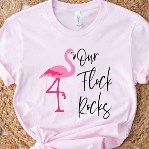 May include: A light pink t-shirt featuring a pink flamingo graphic and the text "Our Flock Rocks" in black script. The shirt is knotted at the bottom and is displayed on a woven, beige rug.