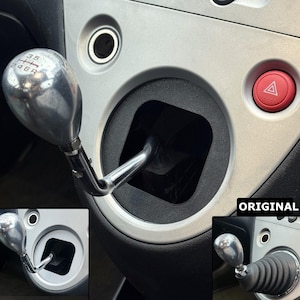 Puede incluir: Un pomo cromado de la palanca de cambios con una bota negra, montado en una consola plateada. El pomo tiene un patrón de 5 velocidades. A la derecha se ve un botón rojo de luces de emergencia.