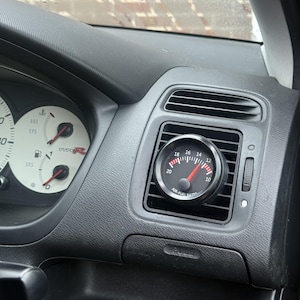 Peut inclure: Un manomètre de rapport air-carburant noir installé dans une bouche d'aération de tableau de bord de voiture. Le cadran noir affiche des chiffres blancs et une aiguille rouge. Le texte "AIR-FUEL RATIO" est visible.