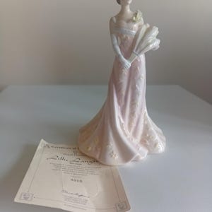Puede incluir: Una figura de porcelana de una mujer con un vestido largo de color rosa pálido con detalles florales blancos, sosteniendo un abanico blanco. Junto a la figura hay un certificado de autenticidad en papel para el modelo Royal Doulton Lillie Langtry HN 3820.
