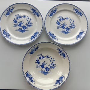 May include: Three round ceramic plates with a white base and a blue floral pattern. Each plate features a central motif of stylised flowers and wheat, with matching floral clusters arranged symmetrically around the outer rim of the plate.