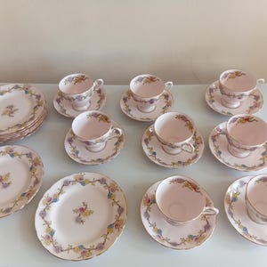 May include: A tea set consisting of six pale pink teacups and saucers, plus three matching plates. Each piece features a delicate floral garland border in blue, yellow, and purple, with a small central floral bouquet on the plates.