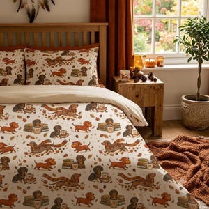 May include: A cream-coloured duvet cover and matching pillowcases featuring a repeating pattern of brown dachshund dogs, stacks of books, mugs, and autumn leaves. The dogs are depicted in various poses, some wearing scarves, interspersed with small acorns.