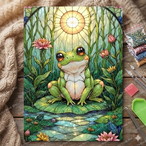 May include: A diamond painting kit featuring a green frog with large eyes sitting on a lily pad in a pond. The scene includes pink lotus flowers, tall green reeds, and a bright circular sun in a stained glass style. Includes tools and beads.