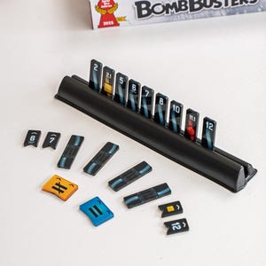 May include: A board game titled "Bomb Busters" with various game pieces. The game includes a black plastic holder with numbered tiles, and several smaller tiles in yellow, blue, and black. The numbers on the tiles range from 2 to 12.