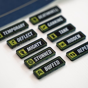Può includere: Una collezione di gettoni di gioco rettangolari neri e verdi con testo bianco. I gettoni presentano parole come "MIGHTY", "STUNNED" e "BUFFED". I gettoni sono disposti su una superficie bianca.
