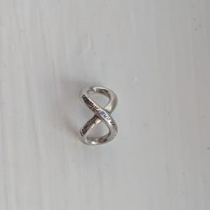 May include: A silver-toned metal charm shaped like an infinity symbol. The surface of the charm features engraved text that reads "Forever and Always" in a cursive script, following the continuous loop of the figure-eight design.