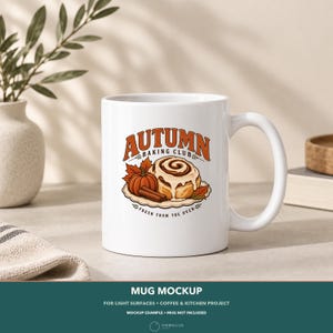 May include: A white ceramic mug featuring a graphic illustration of a cinnamon roll, a pumpkin, and cinnamon sticks. The text reads AUTUMN BAKING CLUB and FRESH FROM THE OVEN in orange and brown lettering, surrounded by small autumn leaf accents.